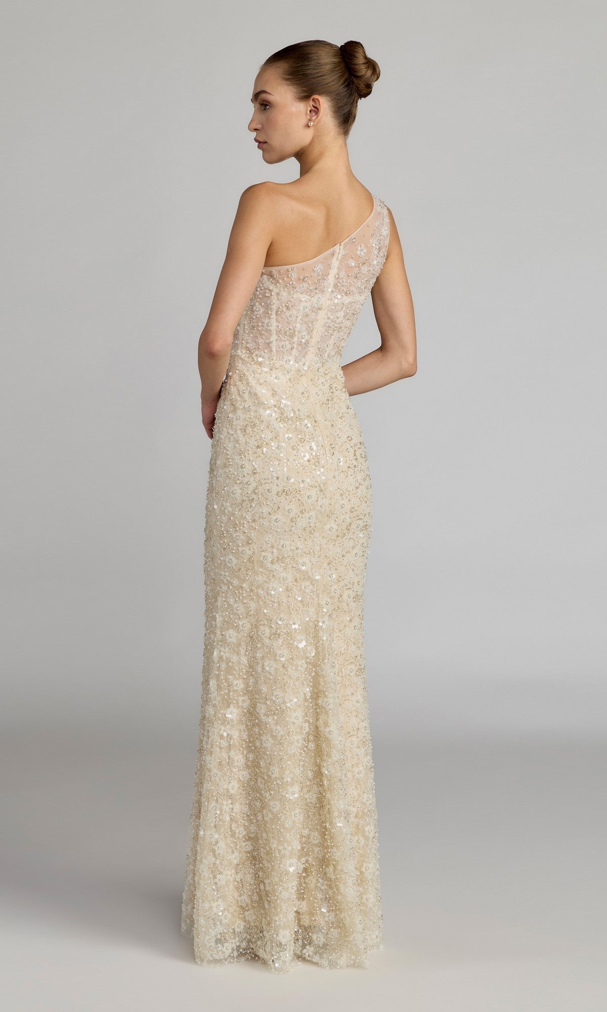 A woman stands with her back to the camera, wearing the Mac Duggal 21287 long formal dress—a cream, sheer one-shoulder gown adorned with delicate sequin embellishments. Her hair is styled in a neat bun, set against a neutral background.